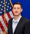 austin cibik with short brown hair and blue collared shirt with black sports coats posing in front of American flag with blue background