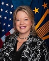 A woman in a black and white printed jacket and black shirt poses in front of the United States and Senior Executive Service flags.