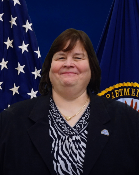 A woman in a black jacket and black and white shirt poses in front of the United States and Department of Veterans Affairs flags.