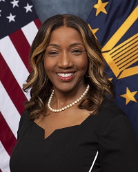 African American woman in a white top and blue blazer poses in front of the U.S. and Senior Executive flags