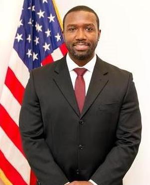 African American male in a grey blazer and white shirt poses in front of U.S. flag